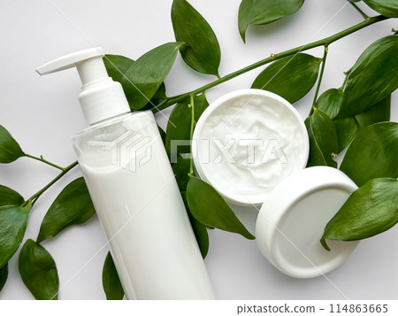 Close up of white lotion bottle and hand cream container with green leaves on white background. Skincare and natural beauty concept. For healthcare, wellness, and organic product poster. Close up of white lotion bottle and hand cream container with green leaves on white background. Skincare and natural beauty concept. For healthcare, wellness, and organic product poster. 114863665
