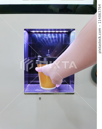 One hand is holding a yellow paper cup at the window of a coin-operated coffee machine. 114863764