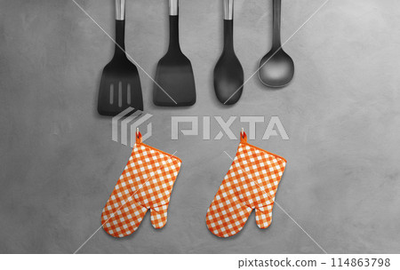 Orange heat resistant cooking gloves with kitchen utensils hanging on cement wall. 114863798