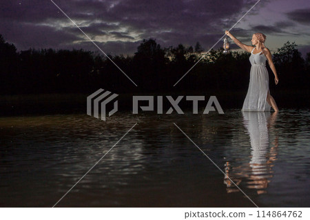 A woman in a flowing dress holds a lantern, walking through water at sunset in a serene scene A woman in a flowing dress holds a lantern, walking through water at sunset in a serene scene 114864762