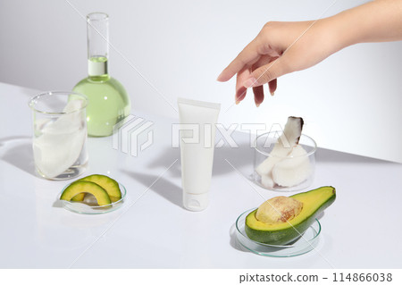 Female hand model is about to pick up the beauty tube. Unbranded tube arranged on the table with few laboratory glassware containing avocados and coconuts. Skincare concept with empty label 114866038