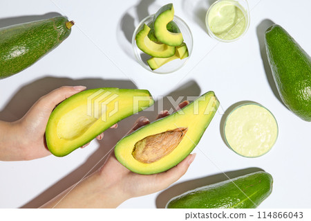 Top view of hand model holding two halves of avocado on white background. Petri dishes of avocado slices and avocado cream displayed. Avocado is very good for skin and hair 114866043