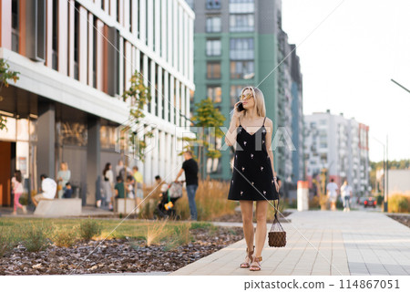 Young woman in a black dress, talking on a cell phone, walking on a city street in the summertime 114867051