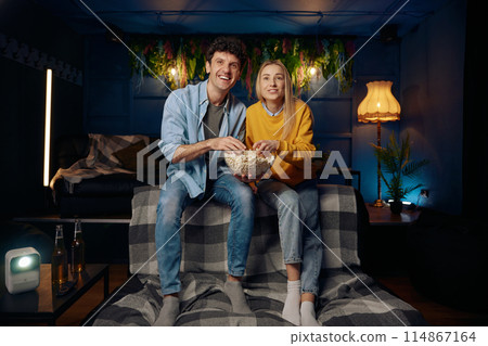 A man and a woman are sitting on a couch, smiling and enjoying popcorn 114867164