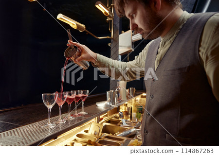 Bartender pouring freshly prepared cocktail into several glasses Bartender pouring freshly prepared cocktail into several glasses 114867263