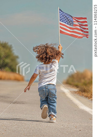 Child running and waving American flag on a rural road in daytime. 114868255