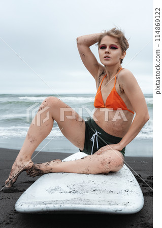 Woman surfer sitting on surfboard on black sand beach with backdrop of ocean waves during summer holiday. Woman looking at camera with confident and sexy smile. She's wearing bikini top and shorts Woman surfer sitting on surfboard on black sand beach with backdrop of ocean waves during summer holiday. Woman looking at camera with confident and sexy smile. She's wearing bikini top and shorts 114869122