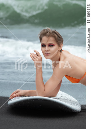 Sexy surfer lying on black sandy beach with surfboard, showcasing fit and athletic body. Caucasian sportswoman is relaxing after sports training session. Sports activities, joy they bring to our lives Sexy surfer lying on black sandy beach with surfboard, showcasing fit and athletic body. Caucasian sportswoman is relaxing after sports training session. Sports activities, joy they bring to our lives 114869138