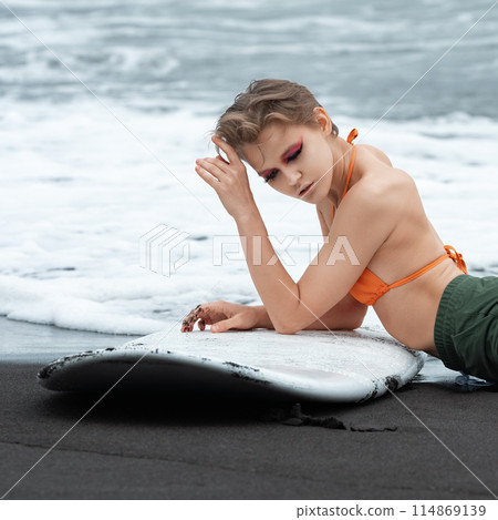 Woman surfer lying on beach with surfboard in relaxed position. Woman's sexuality is suggested by her relaxed pose. Woman who has just finished sports training session during summer vacation on beach Woman surfer lying on beach with surfboard in relaxed position. Woman's sexuality is suggested by her relaxed pose. Woman who has just finished sports training session during summer vacation on beach 114869139