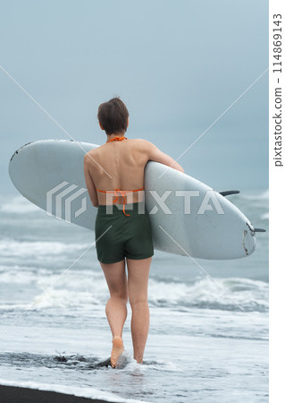 Rear view of female surfer on summer beach carrying her board and looking like an unstoppable force as she strides along sand. It's great picture of woman enjoying sports training during beach holiday Rear view of female surfer on summer beach carrying her board and looking like an unstoppable force as she strides along sand. It's great picture of woman enjoying sports training during beach holiday 114869143