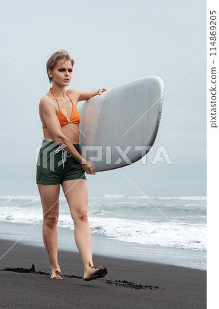 Lovely lady is sportswoman and surfer. She's wearing bikini top, shorts, and is barefoot as she walks on black sand beach, carrying white surfboard against backdrop of sea waves during summer vacation 114869205
