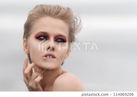 Emotionally stressed woman with bright makeup strikes puzzled pose, pursing her lower lip, raising palm to neck and looking away. Soft selective focus, natural white background and plenty copy space 114869206