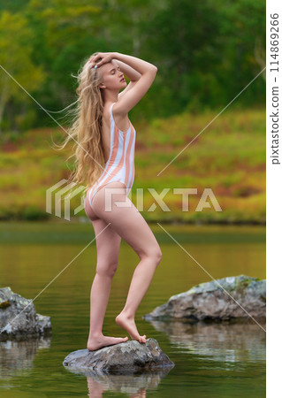 Woman with eyes closed in one piece swimsuit standing on stone rock island in middle of lake surrounded by autumn forest. Sensuality young woman looks so relaxed and happy, enjoying beauty of nature 114869266