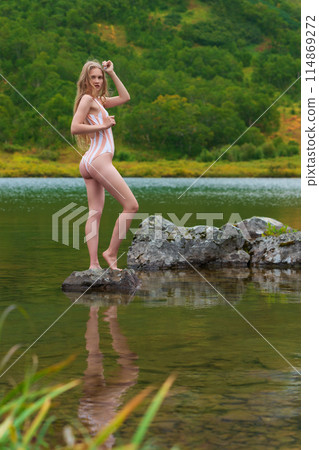 Serene woman in one piece swimsuit with long blonde hair is standing on rocky island in lake, surrounded by colorful autumn forest. Beautiful young Caucasian woman is tourist, relaxing in nature Serene woman in one piece swimsuit with long blonde hair is standing on rocky island in lake, surrounded by colorful autumn forest. Beautiful young Caucasian woman is tourist, relaxing in nature 114869272