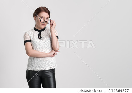 Formal portrait of bossy businesswoman looking at camera over eyeglasses, which she holds with one hand near face. Caucasian business person wearing blouse and trousers, posing on white background 114869277