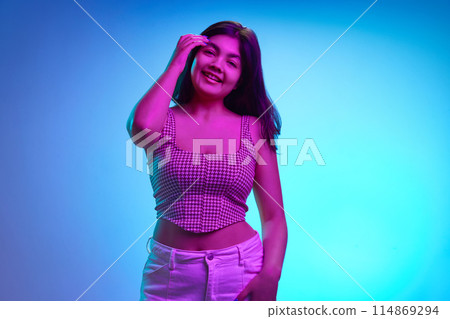 Portrait of young Indian woman looking at camera and cheerful smiling in pink neon light against gradient blue background. 114869294