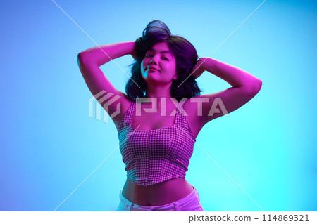 Portrait of beautiful Indian woman posing raising hands in pink neon light against gradient blue background. 114869321