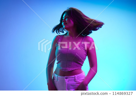 Portrait of charming young Indian girl dancing and her hair fluttering in wind in pink neon light against gradient blue background. Portrait of charming young Indian girl dancing and her hair fluttering in wind in pink neon light against gradient blue background. 114869327