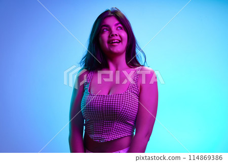 Young Indian girl, 20s years old, looking away with playful expression in pink neon light against gradient blue background. Young Indian girl, 20s years old, looking away with playful expression in pink neon light against gradient blue background. 114869386