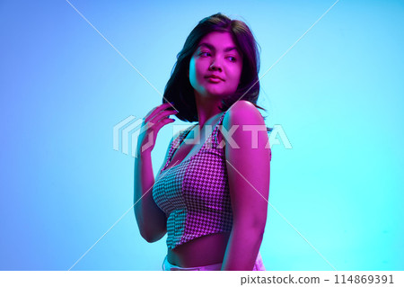 Charming young Indian girl, 20s years old, looking away with playful expression in pink neon light against gradient blue background. Charming young Indian girl, 20s years old, looking away with playful expression in pink neon light against gradient blue background. 114869391