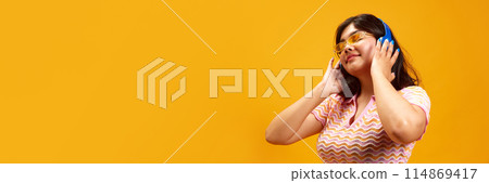 Banner. Indian girl listening music in headphones against vibrant yellow studio background with negative space to insert text. Banner. Indian girl listening music in headphones against vibrant yellow studio background with negative space to insert text. 114869417