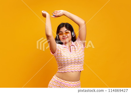 Portrait of charming, active brunette woman dancing while listening music in headphones against vibrant yellow studio background. 114869427