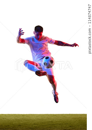 Dynamic shot of young guy dressed sportswear kicking ball in motion in neon light against white studio background. Dynamic shot of young guy dressed sportswear kicking ball in motion in neon light against white studio background. 114874747