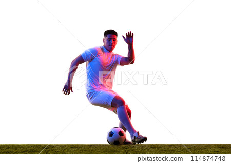Young athletic man training dribbling technique in motion on green lash field in neon light against white studio background. Young athletic man training dribbling technique in motion on green lash field in neon light against white studio background. 114874748