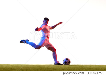 Soccer player in green shorts and white jersey, captured mid-kick on green grass field in neon light against white studio background. 114874753