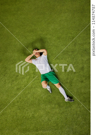 Aerial view of soccer player in green shorts and white jersey lying on grass, covering his face with his hands. 114874787