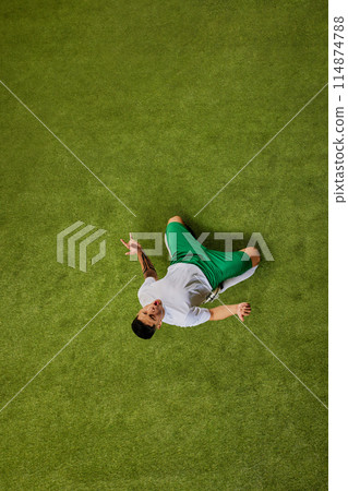 Moment of triumph or celebration. Aerial view of soccer player seated on green grass and gesturing radiating excitement. 114874788