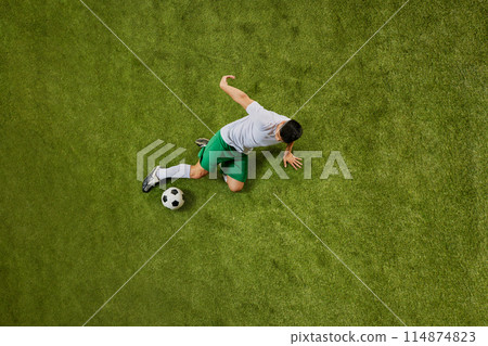 Overhead image of athlete in green shorts and white jersey striking soccer ball with precision on green turf. 114874823