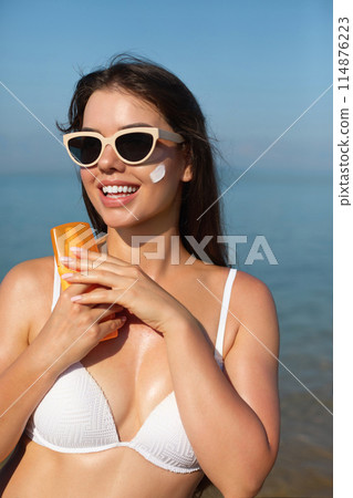 Beautiful woman applying cream sunscreen on a tanned face. Sunscreen. Skin and body care. The girl uses a sunscreen for her skin. Portrait of a female holding suntan lotion and moisturizing sunscreen. Beautiful woman applying cream sunscreen on a tanned face. Sunscreen. Skin and body care. The girl uses a sunscreen for her skin. Portrait of a female holding suntan lotion and moisturizing sunscreen. 114876223