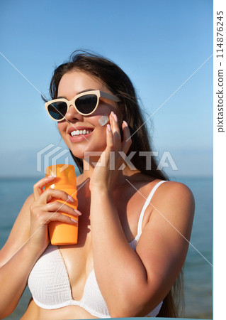 Beautiful woman applying cream sunscreen on tanned face. Sunscreen. Skin and body care. The girl uses a sunscreen for her skin. Portrait of a female holding suntan lotion and moisturizing sunscreen. Beautiful woman applying cream sunscreen on tanned face. Sunscreen. Skin and body care. The girl uses a sunscreen for her skin. Portrait of a female holding suntan lotion and moisturizing sunscreen. 114876245