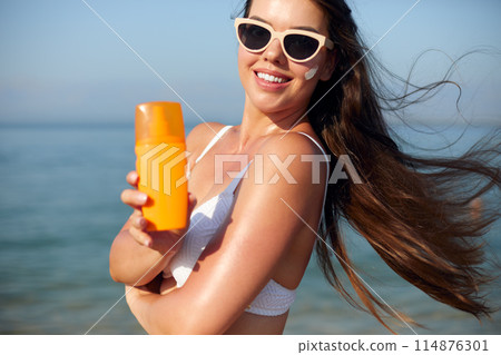 Beautiful woman applying cream sunscreen on a tanned face. Sunscreen. Skin and body care. The girl uses a sunscreen for her skin. Portrait of a female holding suntan lotion and moisturizing sunscreen. Beautiful woman applying cream sunscreen on a tanned face. Sunscreen. Skin and body care. The girl uses a sunscreen for her skin. Portrait of a female holding suntan lotion and moisturizing sunscreen. 114876301