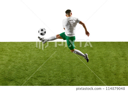 Dynamic photo of young athlete man, soccer player training to kicking ball in mid-air on green field against white studio background. 114879048