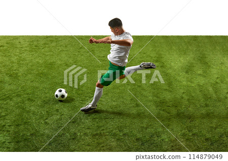 Action shot of young athletic guy, footballer in green and white gear, executing powerful kick on grassy pitch isolated white backdrop. 114879049