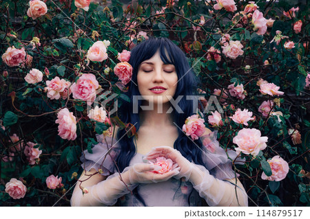 Fairy tale woman portrait. Young beautiful woman Fairy holds pink flower rose on blooming bush background. Hairstyle loose green hair, romantic purple medieval vintage dress. Summer fantasy garden Fairy tale woman portrait. Young beautiful woman Fairy holds pink flower rose on blooming bush background. Hairstyle loose green hair, romantic purple medieval vintage dress. Summer fantasy garden 114879517