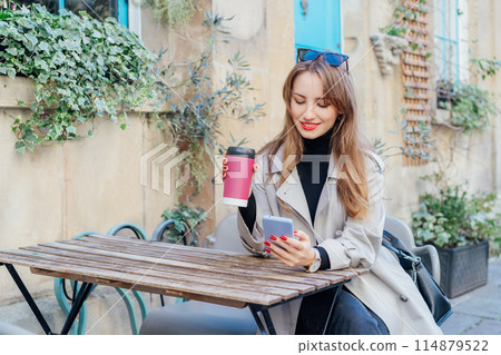 Elegant young woman using phone and drink coffee from reusable cup sitting outdoors. Texting resting in cafe terrace table after city walk. using internet in wifi zone, enjoying coffee and leisure. Elegant young woman using phone and drink coffee from reusable cup sitting outdoors. Texting resting in cafe terrace table after city walk. using internet in wifi zone, enjoying coffee and leisure. 114879522