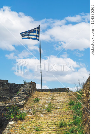 Greek flag waving on a must against sky with clouds on top of Agia Mavra Fort in Lefkada Ionian Island, Greece. 114880363
