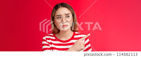 Disappointed doubtful frowning asian girl asking question reassure, looking pointing left displeased, unimpressed with strange place, dislike interiour sulking distressed, stand red background 114882113