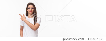 Waist-up portrait of moody, disappointed and complaining cute brunette woman in glasses, frowning and grimacing, sulking defensive, pointing finger upper left corner at something offensive Waist-up portrait of moody, disappointed and complaining cute brunette woman in glasses, frowning and grimacing, sulking defensive, pointing finger upper left corner at something offensive 114882133