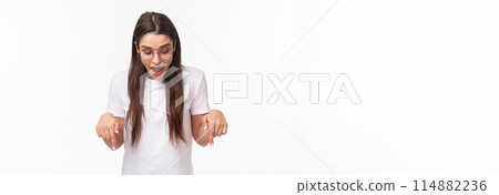 Portrait of amazed, curious happy brunette woman in t-shirt, glasses, looking and pointing down amused, see something awesome at bottom promo, standing white background Portrait of amazed, curious happy brunette woman in t-shirt, glasses, looking and pointing down amused, see something awesome at bottom promo, standing white background 114882236