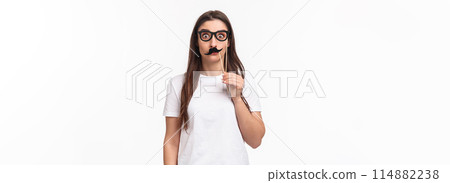 Entertainment, fun and holidays concept. Portrait of funny, playful and carefree girl holding carnaval mask sticks, fancy moustache and glasses, staring camera with popped eyes, white background Entertainment, fun and holidays concept. Portrait of funny, playful and carefree girl holding carnaval mask sticks, fancy moustache and glasses, staring camera with popped eyes, white background 114882238