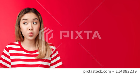 Close-up silly tender young modern asian girl blond haircut hold breath, folding lips and pouting look away as if hiding secretn, stand red background innocent clueless seal lips with promise 114882239