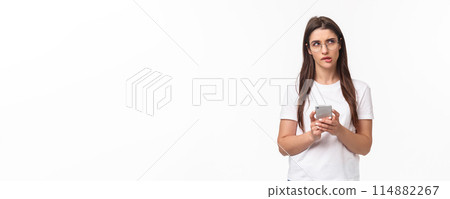 Communication, technology and lifestyle concept. Portrait of thoughtful young woman working remote, answer clients on phone, hold smartphone look up thinking what text back Communication, technology and lifestyle concept. Portrait of thoughtful young woman working remote, answer clients on phone, hold smartphone look up thinking what text back 114882267