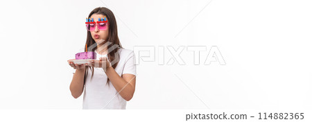 Entertainment, fun and holidays concept. Portrait of cute and funny birthday girl blowing out candle on b-day cake, wear party glasses, making wish for dream come true, white background Entertainment, fun and holidays concept. Portrait of cute and funny birthday girl blowing out candle on b-day cake, wear party glasses, making wish for dream come true, white background 114882365