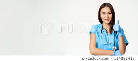 Smiling nurse, asian female doctor in scrubs, showing thumbs up sign and vaccinated arm with medical plaster, recommending vaccination from covid-19, white background Smiling nurse, asian female doctor in scrubs, showing thumbs up sign and vaccinated arm with medical plaster, recommending vaccination from covid-19, white background 114882412