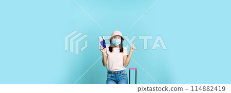 Vertical shot of happy korean woman in medical mask, holding flight tickets and passport, standing with suitcase, dreaming of future vacation, blue background 114882419