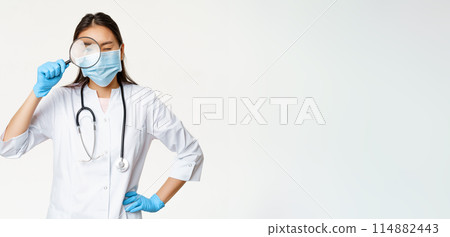 Asian female doctor looking through magnifying glass, examine patient, searching disease, wearing medical mask and rubber gloves, white background 114882443